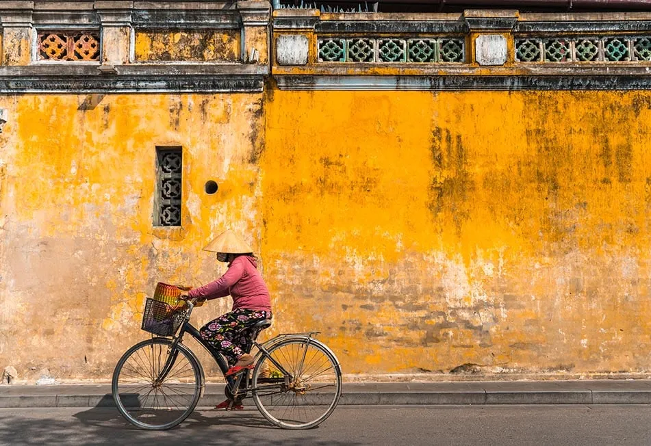 A backpacker on a motorbike wearing a conical hat in a rural Vietnamese village, symbolizing authentic travel and adventure for The Broke Backpacker Vietnam.