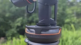 A black and white Celestron Origin amateur astronomy telescope in a field with trees behind it, showing handles for transport
