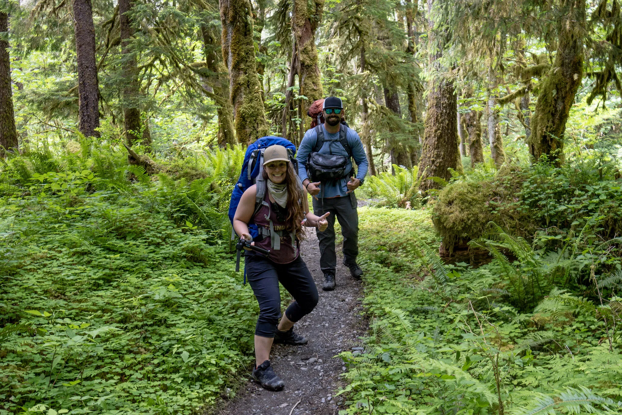 A boyfriend/girlfriend couple stand on a trail in a green forest with overnight packs on. They are seen from behind, walking away down the path surrounded by lush trees.