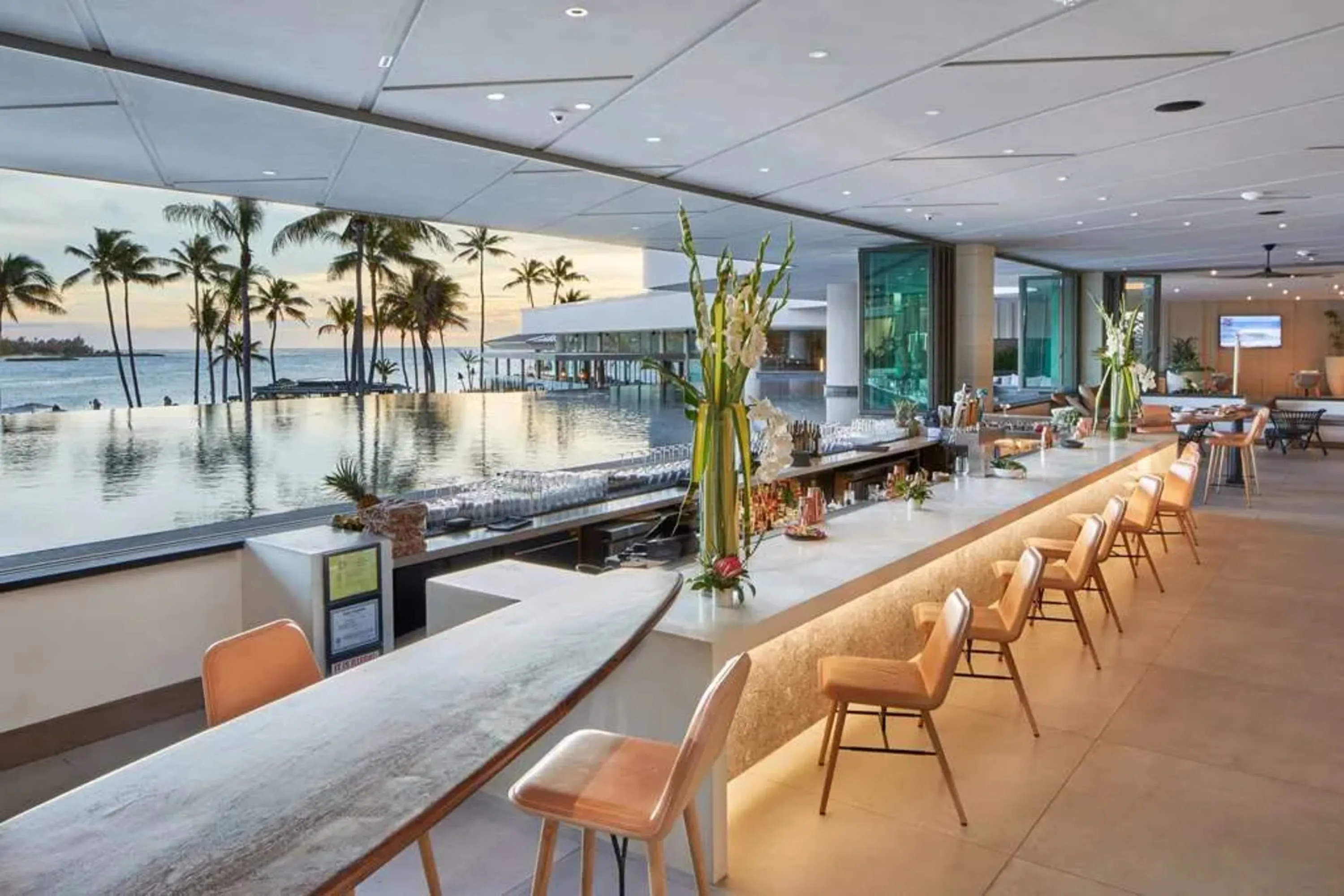 A chic outdoor bar with neon-lit seating. The bar is set right next an infinity pool that overlooks a palm-dotted beach