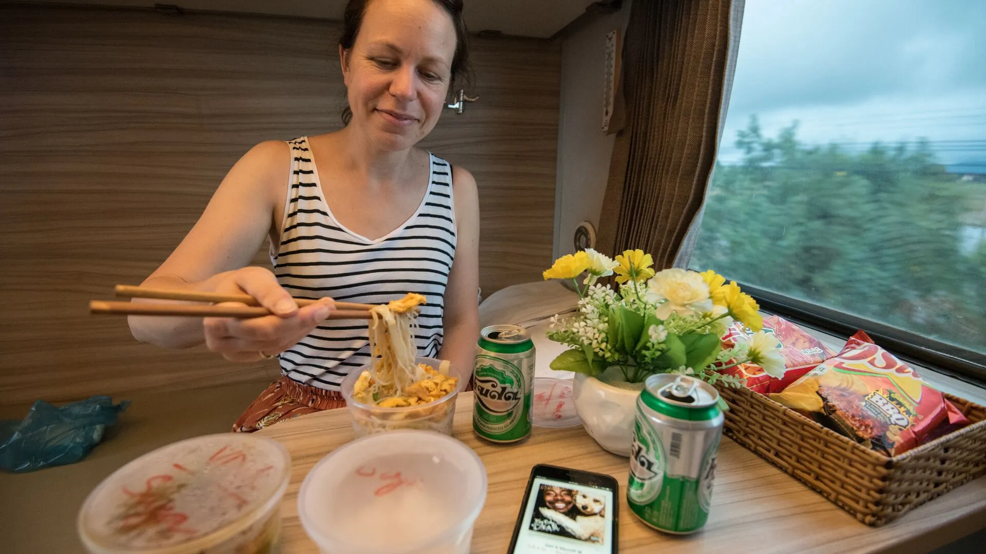 A female traveller enjoys a container of noodles on her train table. Also on the train table are two canned beverages, a few packets of crisps and a small flower pot with yellow flowers.