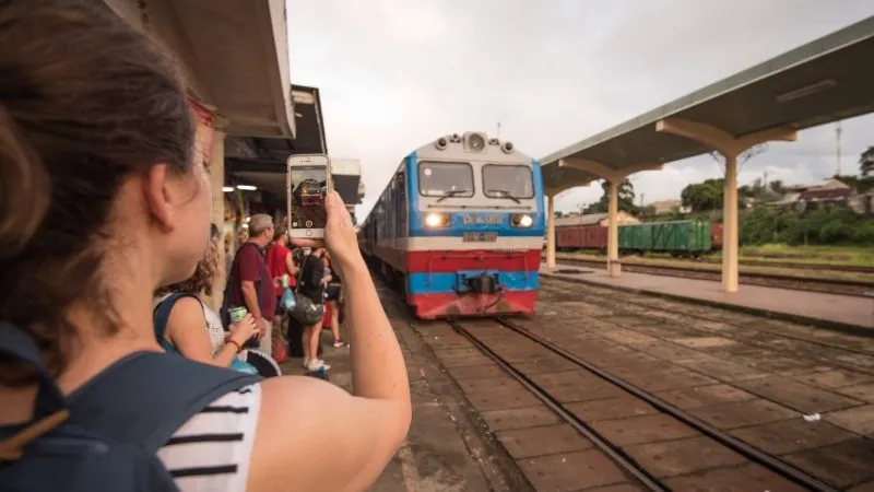 A female traveller holds up an iphone to take a photo of an approaching blue and red train.
