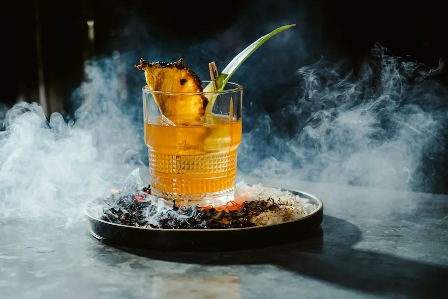 A festive holiday cocktail garnished with pineapple and leaf, served on a smoking plate