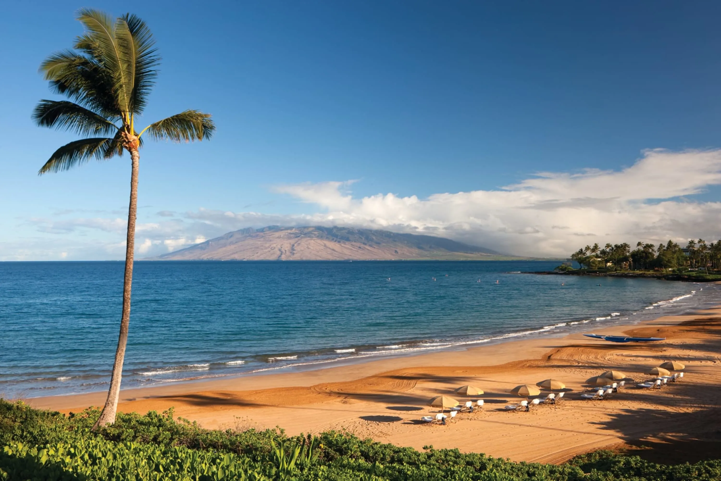 A few loungers sit on a well-manicured beach overlooking calm waters and a large Maui mountain in the distance