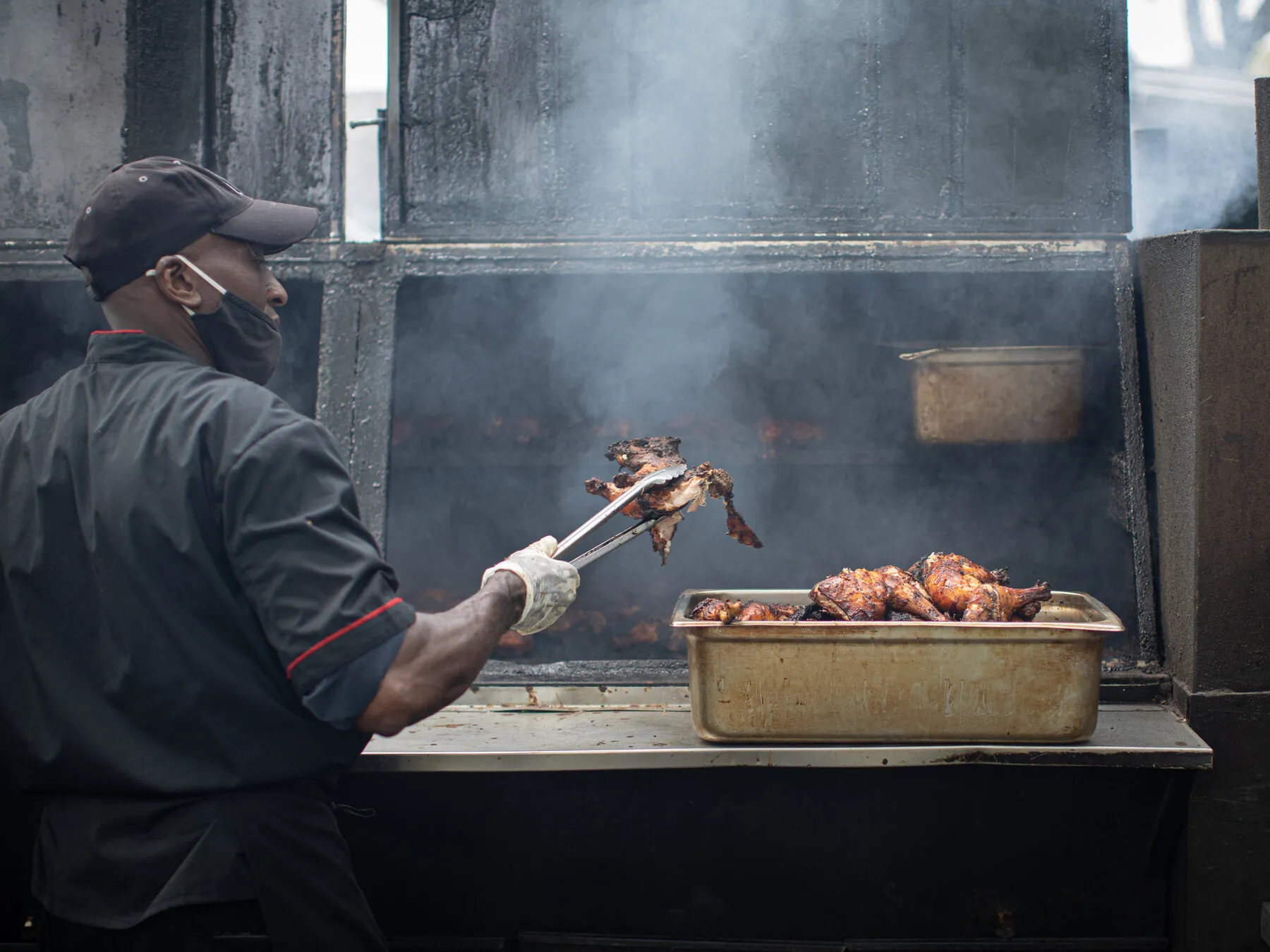 A large outdoor charcoal grill at Forever Jerk, used for cooking jerk chicken and pork, with smoke rising.