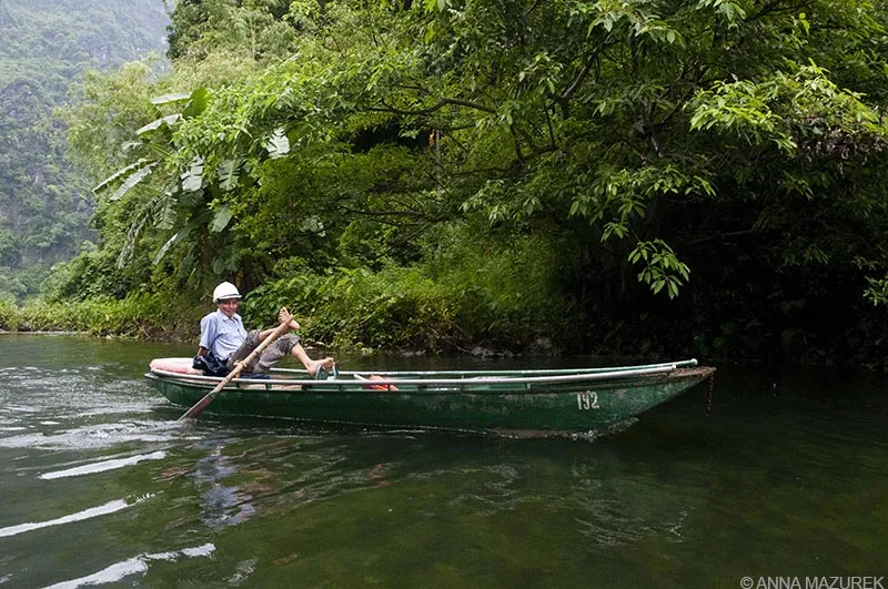 A person rowing a traditional Vietnamese boat with their feet through a cave tunnel, surrounded by lush green plants and rocky formations.