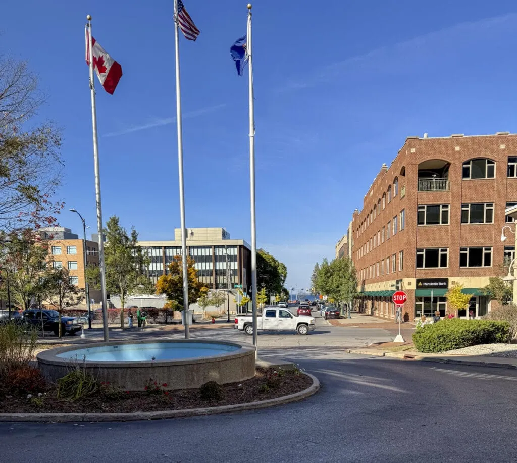 A roundabout with a small fountain and three flagpoles stands in the foreground, just outside the Park Place Hotel in Traverse City, Michigan. Surrounding buildings include a multi-story brick structure and a modern office complex, showcasing this good place to vacation in Michigan. Vehicles, trees, and a clear blue sky complete this picturesque setting.