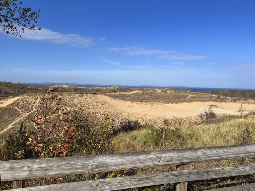 A scenic view of vast sand dunes under a clear blue sky, with sparse vegetation in the foreground. A wooden fence runs across the bottom of the image, reminiscent of the picturesque landscapes found in some of the best Michigan towns on Lake Michigan, with a distant horizon featuring a glimpse of the lake. This demonstrates why it's one of the good places to vacation in Michigan.
