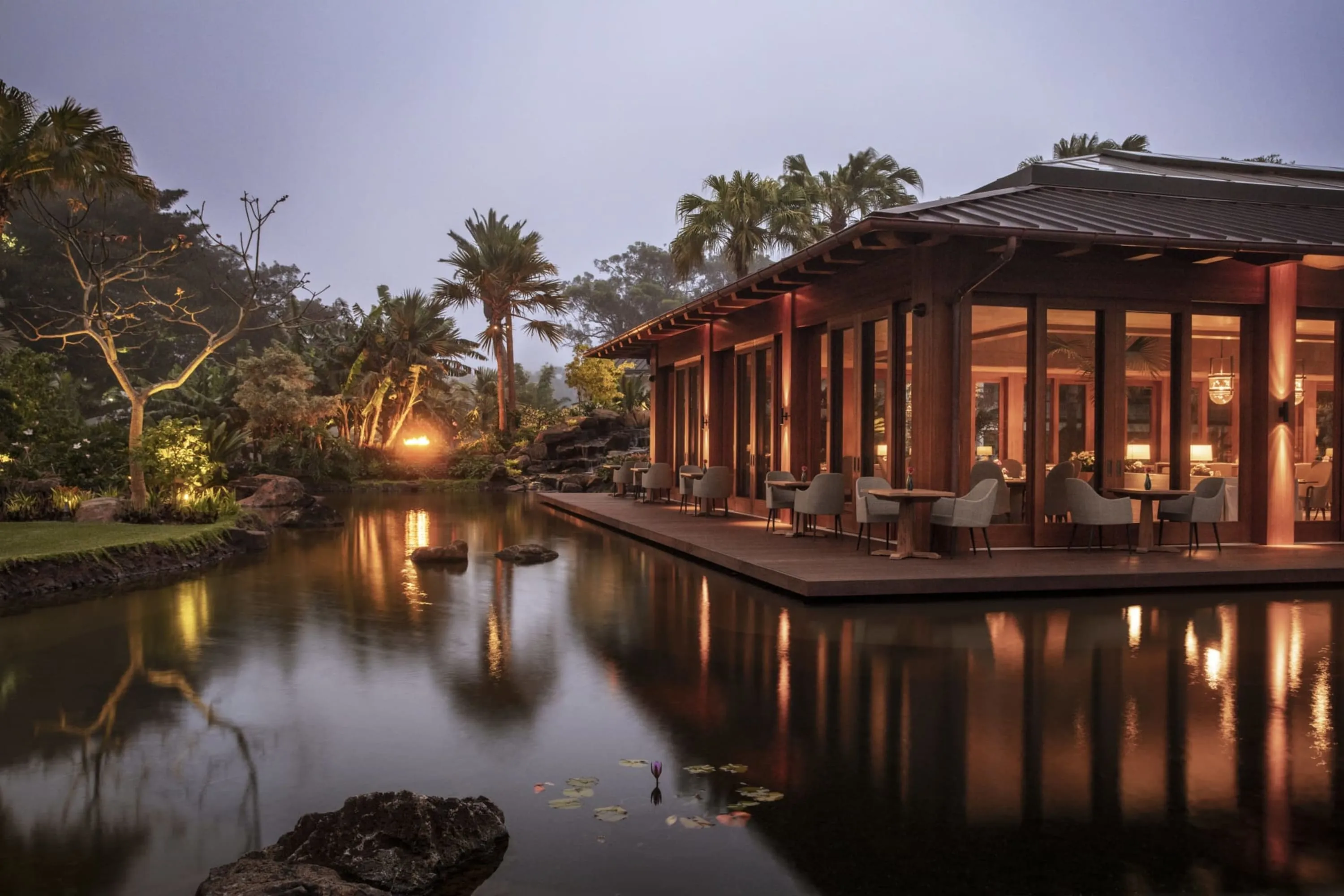 A serene pond borders an upscale eatery with glass and wood walls. In the distance a bonfire is lit within the jungle