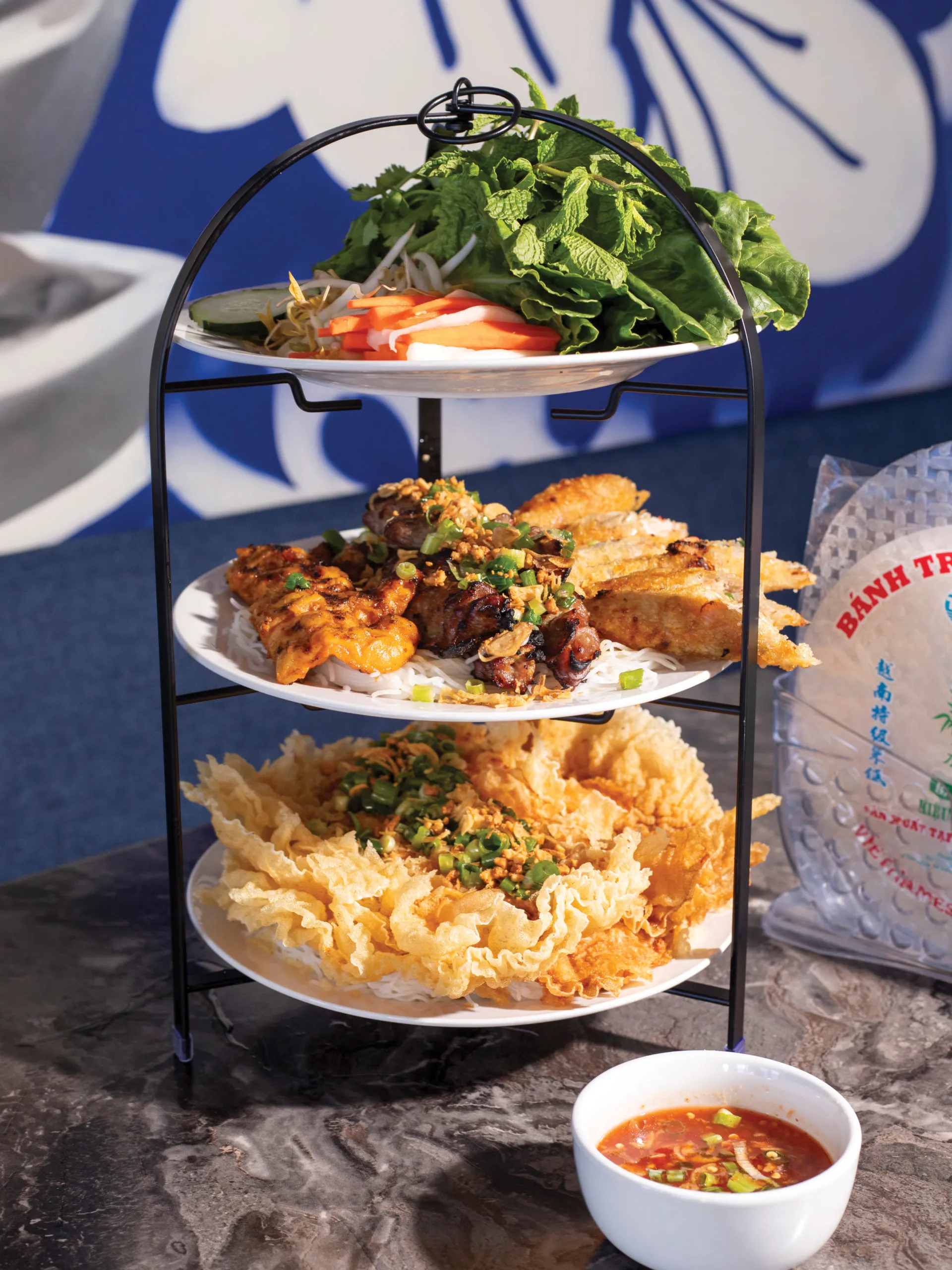 A tiered tower holds various Vietnamese components like pork skewers, fried shrimp paste, noodles, and herbs for making spring rolls.
