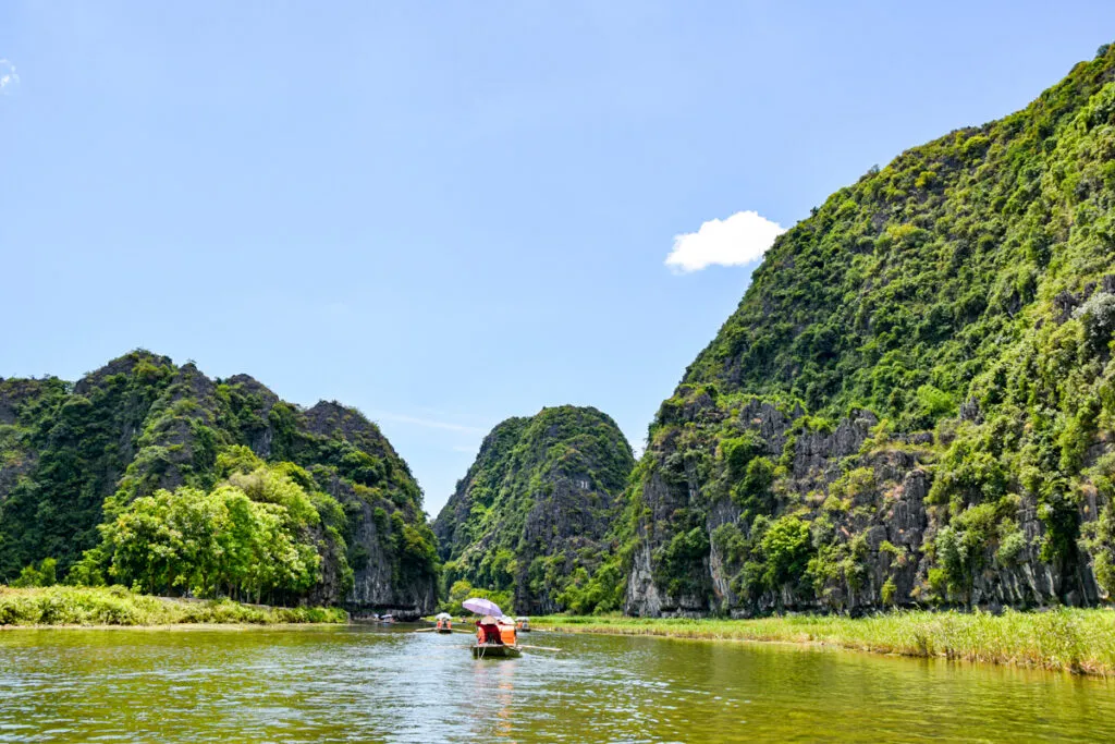 A traditional boat with passengers being rowed down a river lined with limestone karsts and lush vegetation in Tam Coc