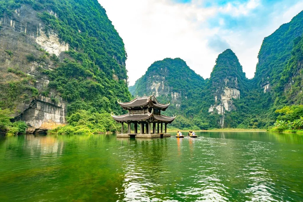 A traditional Vietnamese boat navigates through a scenic waterway surrounded by towering limestone karsts in Trang An