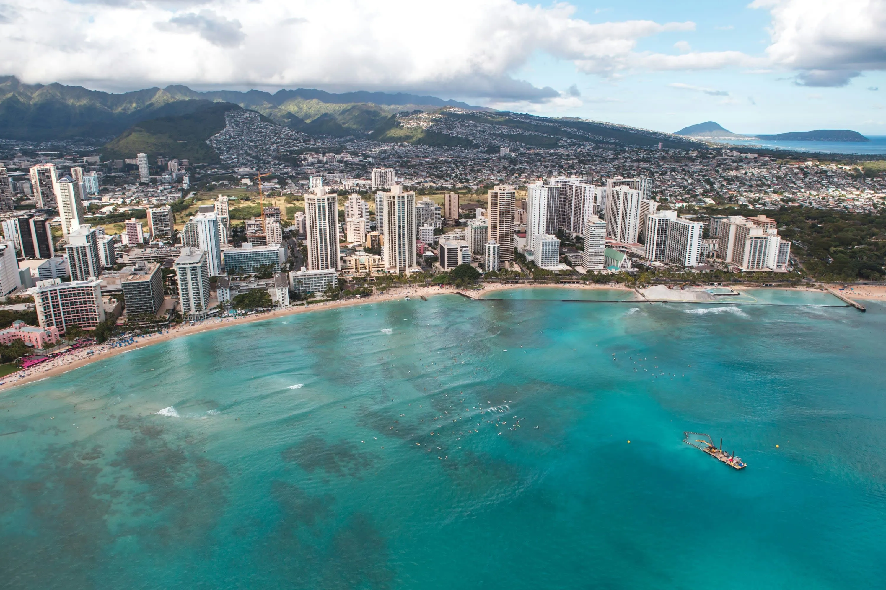 Aerial view: whitewashed skyscrapers line up against turquoise waters with urban sprawl leading up to green mountains in the background