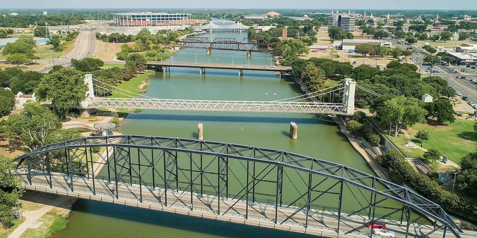 Alt text: A view of the Brazos River flowing through a rural landscape with green banks and trees under a partly cloudy sky.