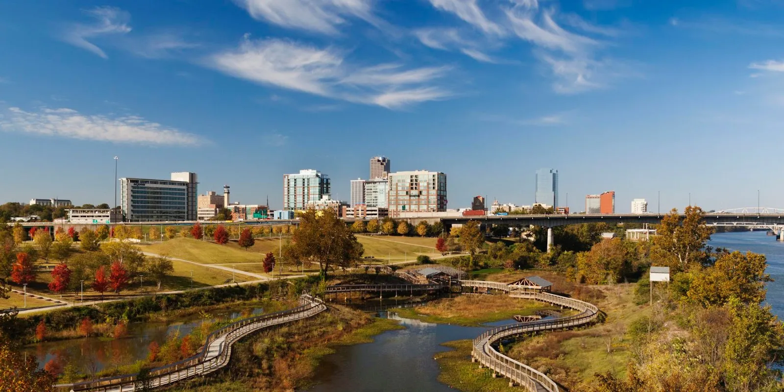 Alt text: The Little Rock skyline viewed from across the Arkansas River, showing modern buildings reflecting in the water under a bridge.