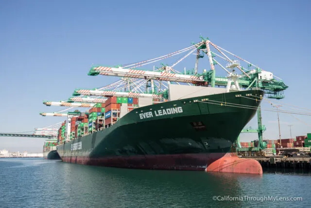Boat cruising in a harbor with large cargo ships and cranes