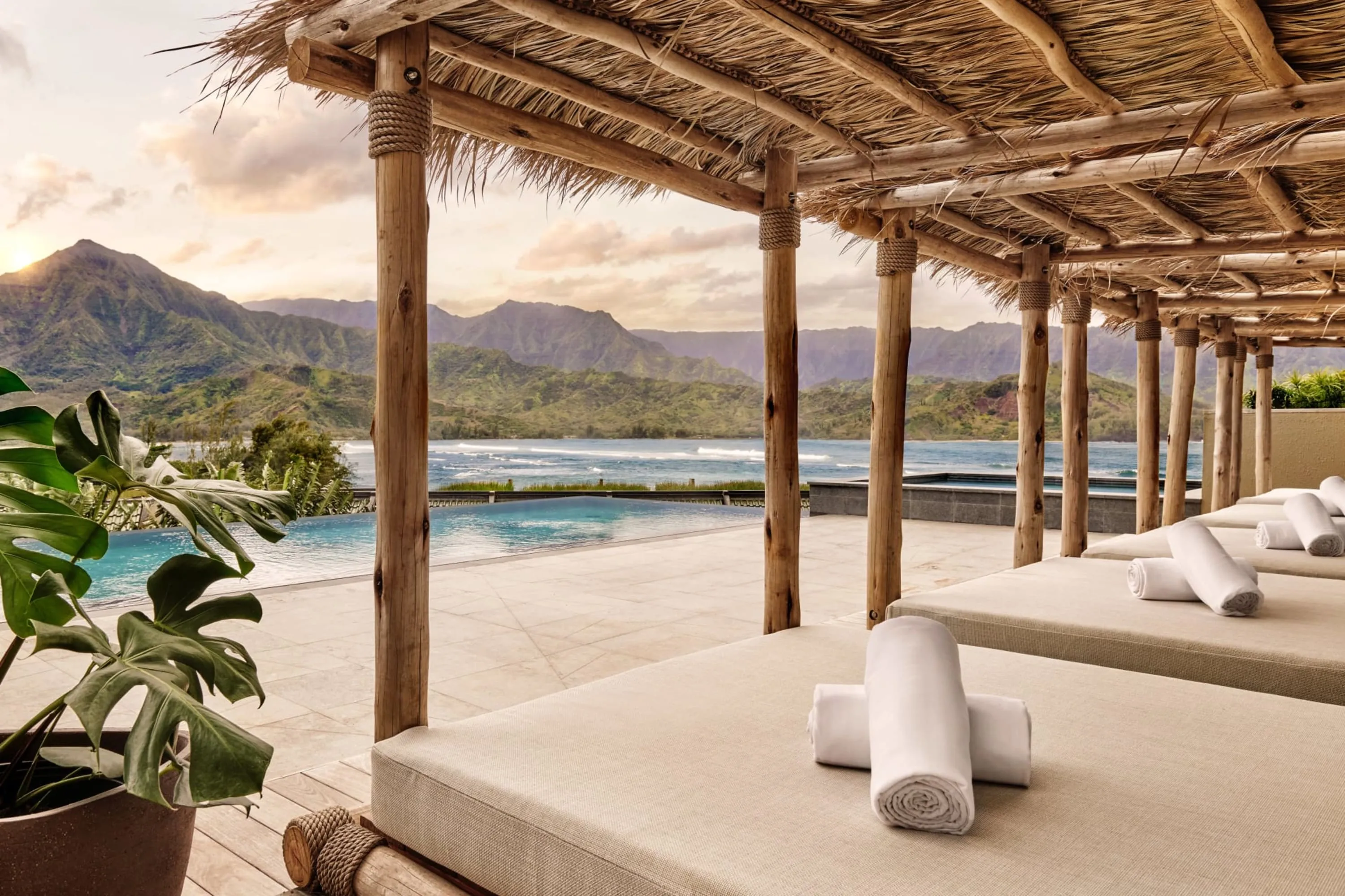 Front and just off-center: a series of luxe lounger beds under thatch awnings. To the left, an infinity pool over looks semi-rough surf with the green mountains of Kauai in the distance