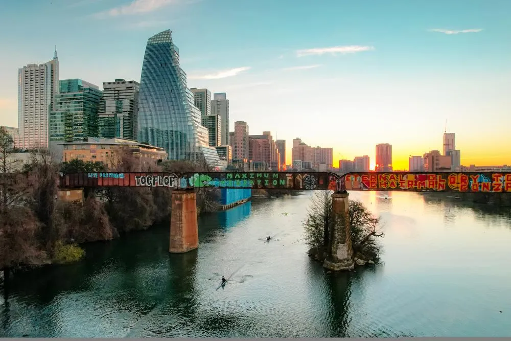 Kayakers paddling under a bridge with grafitti on Lady Bird Lake while the sun sets against Downtown Austin buildings.