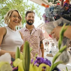 People smiling on a sunny day in front of bunches of flowers at the Portland Saturday Market, a popular spot for fun things to do in Portland