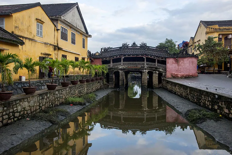 The iconic Japanese Covered Bridge in Hoi An, Vietnam, spanning a small canal with traditional yellow-walled buildings and lanterns visible in the background.