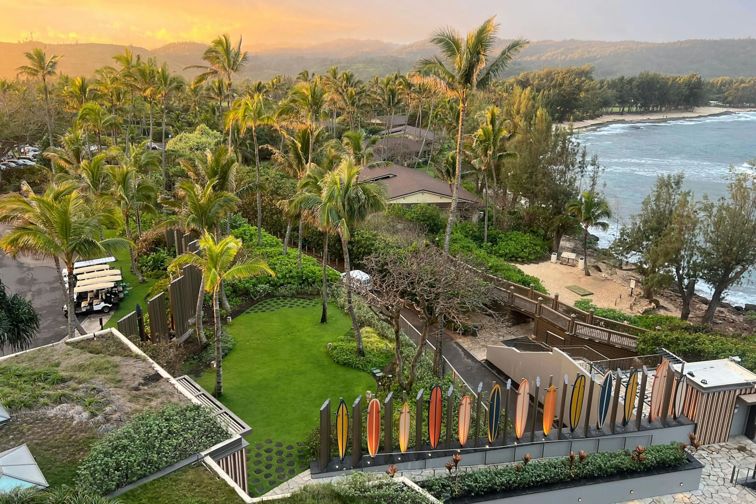 The resort grounds of The Ritz-Carlton O‘ahu, Turtle Bay with the sun setting over the hills in the distance, choppy waves on a lovely private beach and surfboard art 