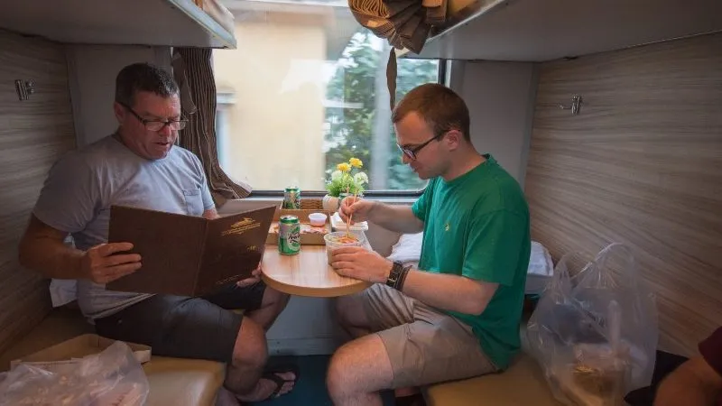 Two travellers enjoy noodles and canned beverages in their train compartment. They