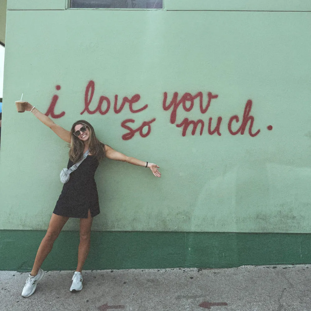 Woman standing with arms wide and iced coffee in hand in front of the iconic I Love You So Much Mural in Austin, Texas.